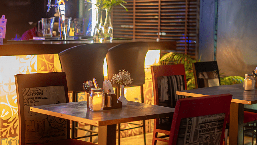 A warmly lit restaurant interior with a bar in the background and tables and chairs in the foreground. The bar has a white counter and hanging lightbulbs. The tables have candles and flowers on them, and the chairs are upholstered in red fabric with newspa